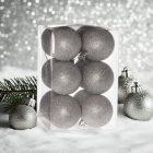 Ezüst glitteres műanyag karácsonyi gömb szett (12×6 cm) átlátszó dobozban, havas felületen, ezüst háttérrel.