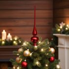 Piros üveg csúcsdísz karácsonyfán, arany és piros gömbökkel, meleg fényű gyertyákkal és fa burkolatú háttérrel.