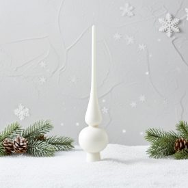 Fehér üveg csúcsdísz havas felületen, fenyőágakkal és tobozokkal, szürke háttér előtt hópelyhekkel.