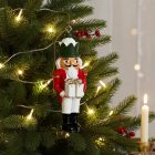 Díszes diótörő katona akasztóval lóg a feldíszített karácsonyfán, gyertyafénnyel a háttérben.