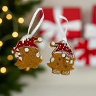 Két mézeskalács gnóm karácsonyfadísz piros sapkával, fehér szalagon, ünnepi fények és piros ajándékok előtt.