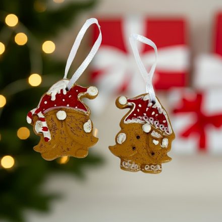 Két mézeskalács gnóm karácsonyfadísz piros sapkával, fehér szalagon, ünnepi fények és piros ajándékok előtt.